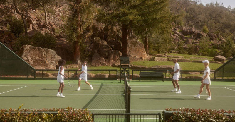 Four people playing tennis on a court with trees and rocks in the background